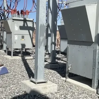 A person wearing a white hard hat and safety glasses holds a clipboard while inspecting equipment at an outdoor electrical substation surrounded by gravel and metal structures.